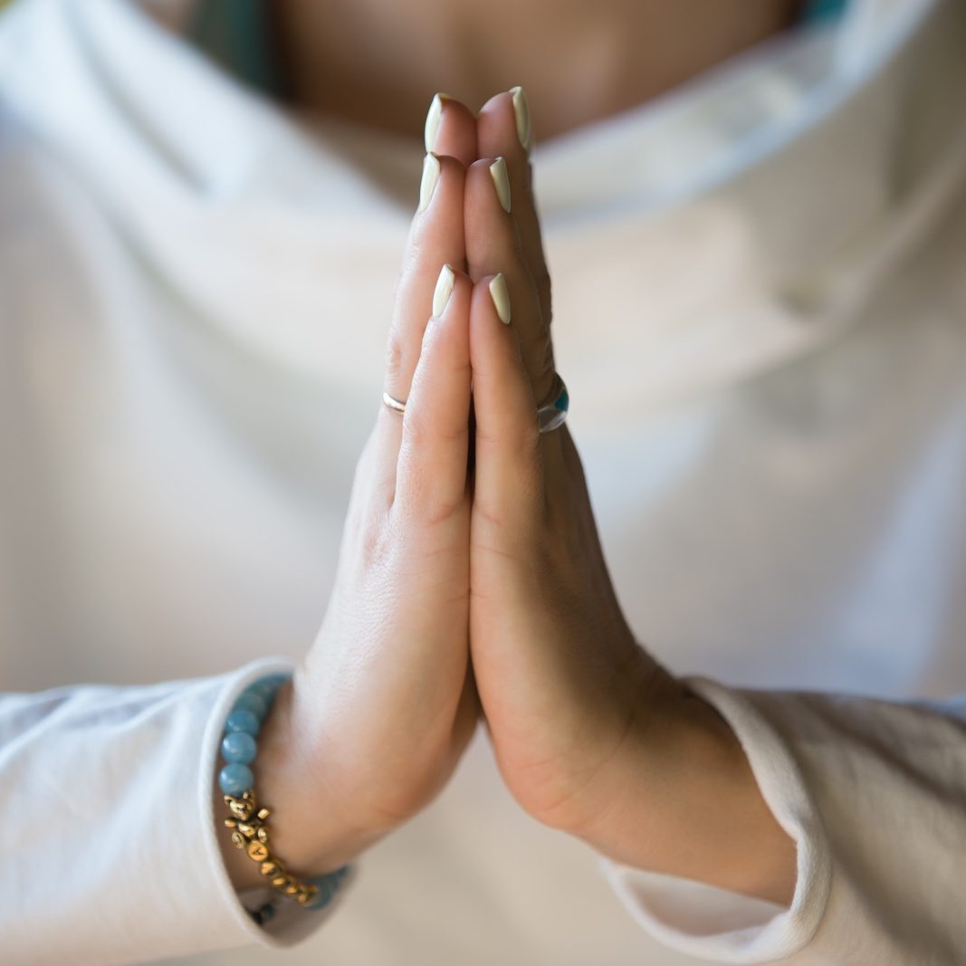 Close-up of hands of sporty young beautiful woman in white clothes meditating indoors, focus on arms in Namaste gesture